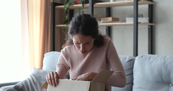 Frustrated upset young woman customer unpacking delivery at home. Angry dissatisfied shopper opening cardboard box feels bad surprise about damaged wrong bad parcel. Postal shipping problem concept