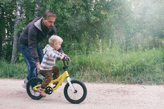 Father Teaches His Little Child To Ride Bike In Spring Summer Park. Happy Family Moments. Time Together Dad And Son. Candid Lifestyle Image.