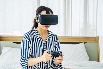 Asia woman wearing VR glasses and sitting in bed in the bedroom. Holding joy stick and playing game by using VR glasses, quarantine due coronavirus pandemic. stay at home..