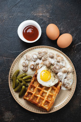Belgian waffle with champignons, fried egg and pickles, flatlay on a dark brown stone background, vertical shot