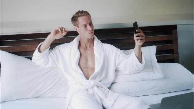 Handsome blond guy in white bathrobe make selfie photo on mobile phone in bed