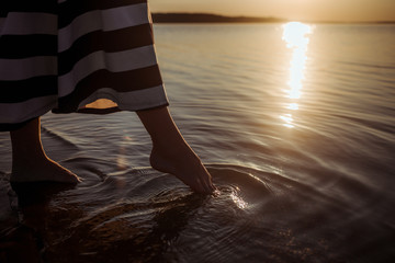 During sunset a girl in a striped dress walks along the beach, only her barefeet in the water are visible. Circles of life diverge on water. The state of calm and pacification.