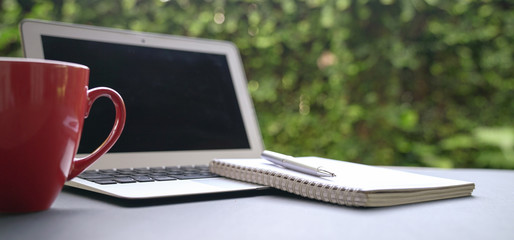 Computer laptop with notepad and coffee in red cup with green nature background. Work remotely or...