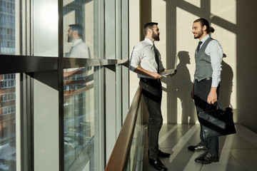 Two young successful businessmen standing by large window during discussion