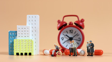 Miniature elderly people lining up in front of alarm clocks and a miniature hospital.