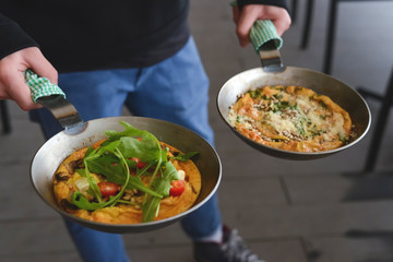 Man holds two pans of English breakfast - fried egg, beans, tomatoes, mushrooms, seeds served in a pan.