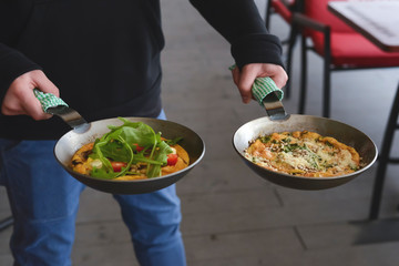 Man holds two pans of English breakfast - fried egg, beans, tomatoes, mushrooms, seeds served in a pan.
