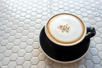 Soft milk foam above the coffee cup with a black saucer on the table with a white hexagon mosaic pattern.