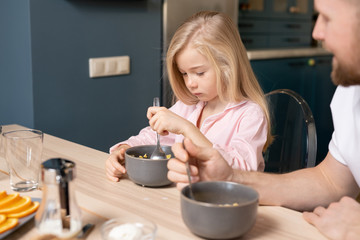 Cute sad girl putting spoon into bowl with muesli while having breakfast