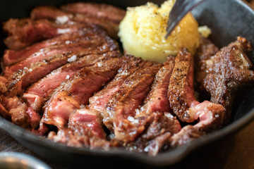 Cast iron plate with sliced grilled rib-eye cooking a medium-rare steak with mashed potato and sauce in saucer over table.