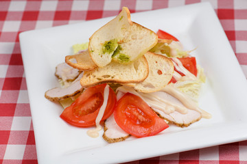 Caesar salad with grilled chicken and fresh tomatoes served on white plate on red plaid background.
