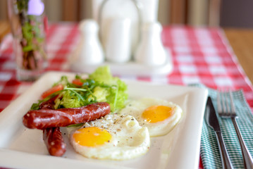 Breakfast with salad, fried egg and sausages served in white plate over red plate tablecloth.