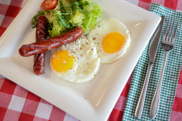 Breakfast with salad, fried egg and sausages served in white plate over red plate tablecloth.