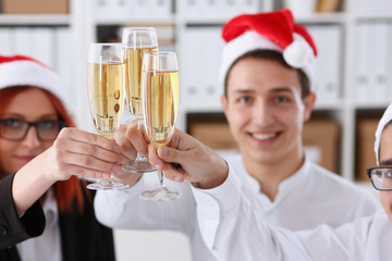 A group of businesspeople celebrating Christmas New Year corporate in the office while toasting holding glasses of champagne in hands on their heads wearing Santa cap.