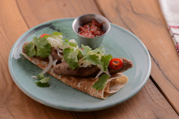 Meat sausages with fresh greenery on lavash bread with sauce in a sauce pan served on a blue plate over wooden table.