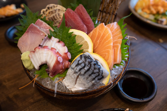 A combination sashimi set consisting of pickled saba, salmon, squid, tuna and hamachi on a dark bowl with ice on the bottom and beautifully decorated. Placed on a dark brown wooden table.