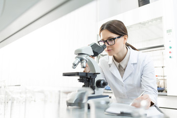 Serious female researcher in whitecoat looking in microscope by workplace