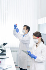 Young female lab worker scrolling in tablet while her colleague looking at flask