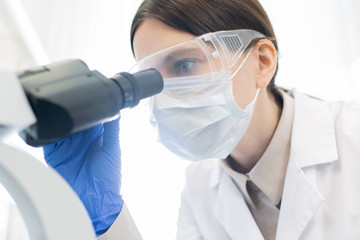 Female researcher in gloves, mask and protective eyewear bending over microscope