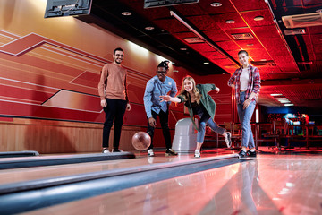 Pretty young female throwing bowling ball on track during play with friends - obrazy, fototapety, plakaty