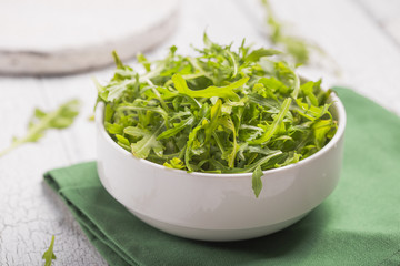 Fresh green arugula leaves on white bowl, rucola rocket salad on wooden rustic background with place for text. Selective focus,  healthy food, diet. Nutrition concept