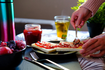 Sweet breakfast with freshly made jam on toast and a very hot tea or honey camomile served by a girl. She also spreads the jam