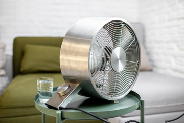 Modern fan with a glass of water on the table at home. Concept of abnormal summer heat and thirst