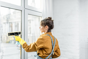 Beautiful young housewife in working clothes washing windows by professional tools at home