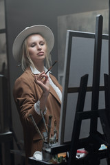 Young elegant woman with paintbrush looking at unfinished painting