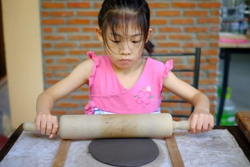 Fototapeta na wymiar A cute Asian preschooler rolling her gray clay using a wooden roller into a flat slab to mold it into a dish into pottery class.