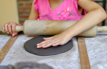 A cute Asian preschooler rolling her gray clay using a wooden roller into a flat slab to mold it into a dish into pottery class.