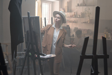 Young serious woman in hat and casualwear standing in front of easel in studio