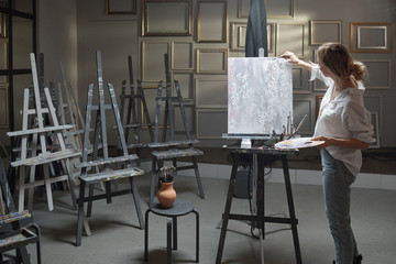 Young blond woman in casualwear standing in front of picture on easel