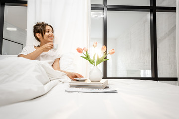 Young and pretty woman enjoying bedtime, lying with coffee cup in the white and sunny bedroom...