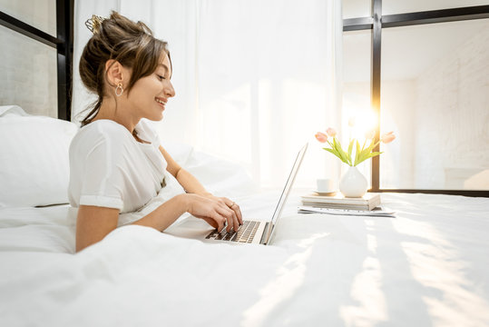 Woman Working On A Laptop, Lying In The Beautiful White And Sunny Bedroom, Wide Interior View With Copy Space Work From Home At Cozy Atmosphere Concept.