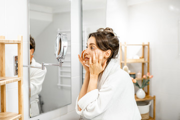 Young woman in bathrobe taking care of her skin, looking at the mirror in the bathroom. Facial skin...