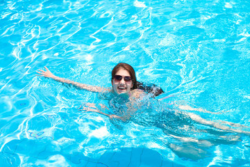 Beautiful joyful girl with long hair in sunglasses swims in the pool on a summer day. Travel, resort