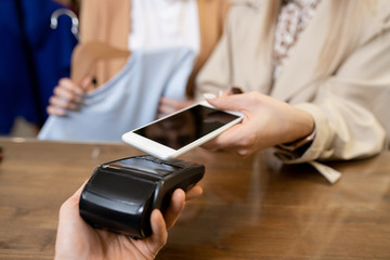 Young consumer holding smartphone over payment machine held by shop assistant