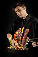 A man in a black shirt prepares food on a dark background. Meat, potatoes, tomatoes, carrots and other vegetables fly in the air. Blurred as an artistic effect. Motion, flight and levitation concept.