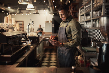 Cheerful young afro-american cafe owner wearing black striped apron using digital tablet - obrazy, fototapety, plakaty