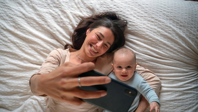 Authentic close up of neo mother and her newborn baby making a selfie or video call to father or relatives in a bed. Concept of technology, new generation,family, connection, parenthood, authenticity