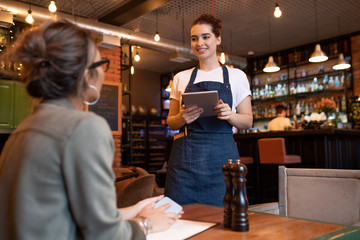 Happy young waitress with digital tablet standing in front of female guest - obrazy, fototapety, plakaty