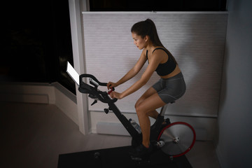 Fototapeta na wymiar Home workout indoor stationary bike Asian girl biking screen with online classes woman training on smart fitness equipment indoors for cycling exercise. Late at night in bedroom.