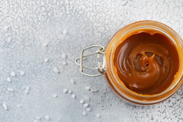 Glass jar with salted caramel and spoon on light background, copy space