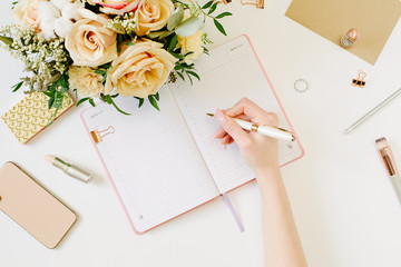 Hand wtite in pink opened notebook on white background. Tender mood and blogging concept with beautiful roses bouquet and accesoires in flat lay style