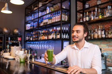 Young smiling barman holding glass of green salary cocktail or smoothie