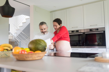 Happy pregnant woman with her husband sitting at home. Smiling couple consulting a book at the kitchen.