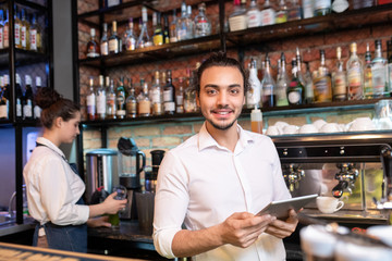 Young cheerful waiter with touchpad standing in front of camera