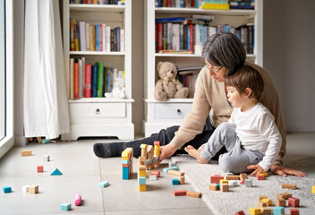 Little toddler boy playing wooden building blocks with grandmother at home. Family spending time together at quarantine period during pandemic. Bright lifestyle.