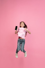 Jumping high with phone. Caucasian little girl portrait isolated on pink studio background. Cute brunette model in shirt. Concept of human emotions, facial expression, sales, ad, childhood. Copyspace.